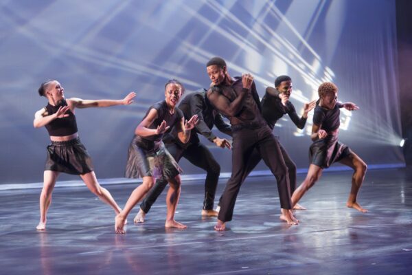 Six dancers dressed in black push and pull against each other on a stage with a blue backdrop and white streaks of light