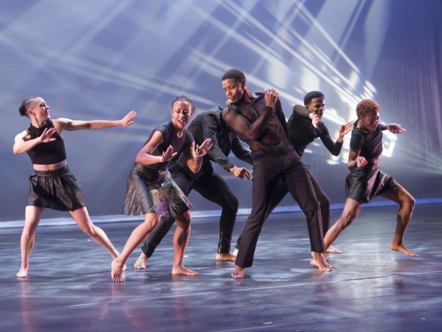 Six dancers dressed in black push and pull against each other on a stage with a blue backdrop and white streaks of light