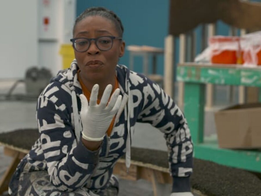 A woman with medium-brown toned skin wearing a black jumpsuit with white lettering on it gestures with her right hand