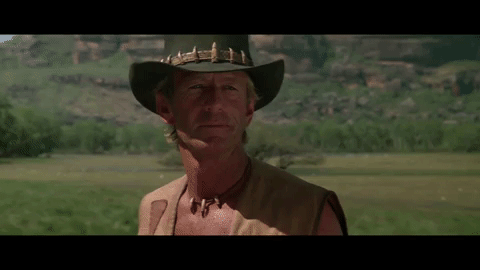 A shirtless Crocodile Dundee stands in a plains area wearing his signature leather hat with teeth