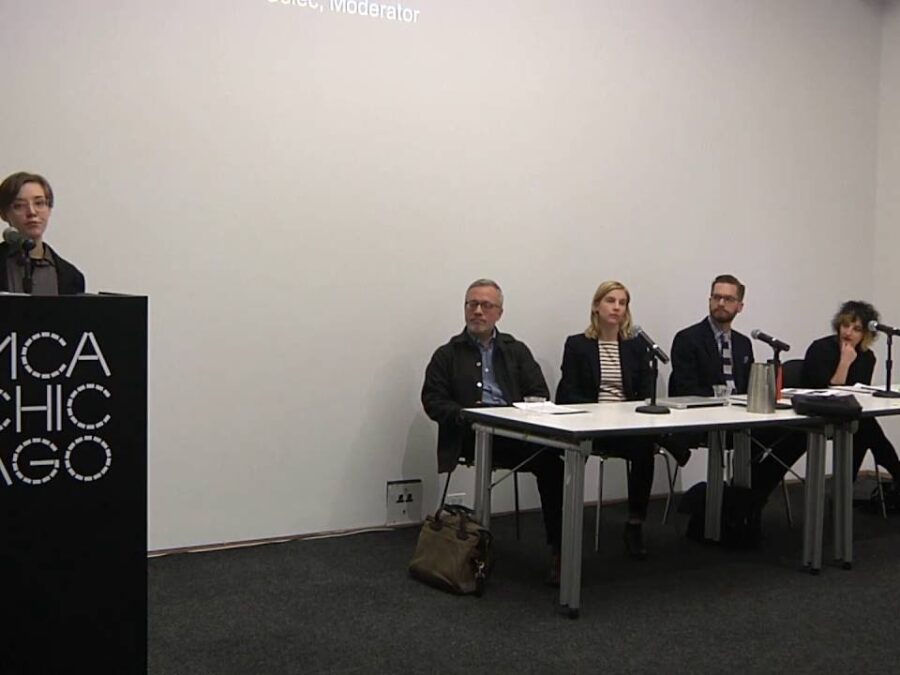 One person stands behind a black podium with the MCA Chicago logo on it as four people sitting at a table look on