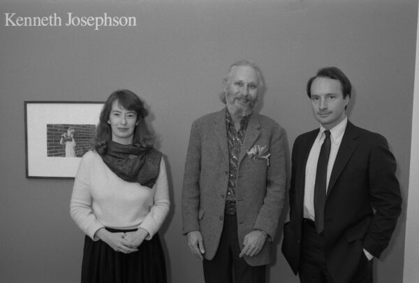 Black and white photo of one woman and two men posing next to a framed photograph with the name Kenneth Josephson on the wall above