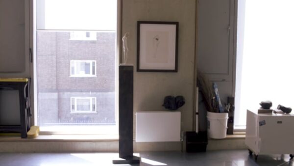 A comfortable modern apartment with a floor to ceiling window with sunlight streaming onto a tall skinny pedestal with a figural sculpture on it