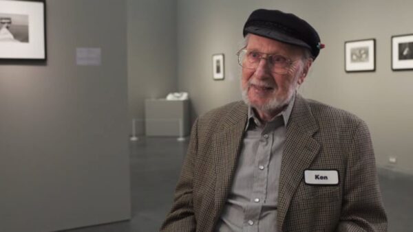 An older man with reddish-peach skin sits in a gallery wearing a cap, suitcoat, and nametag labeled Ken.