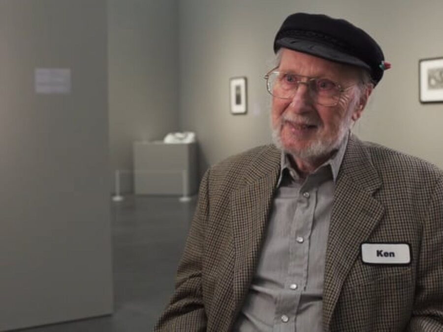 An older man with reddish-peach skin sits in a gallery wearing a cap, suitcoat, and nametag labeled Ken.