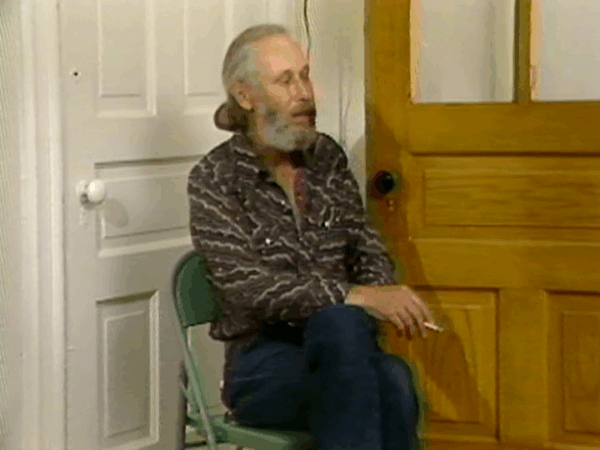 A man with a greying beard and long hair pulled into a bun at the nap of his neck sits near two doors in a corner holding a cigarette in his right hand