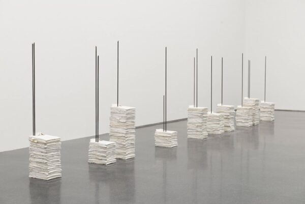 Eight piles of white shirts of various heights with rebar pierced through them in a row in a white gallery space
