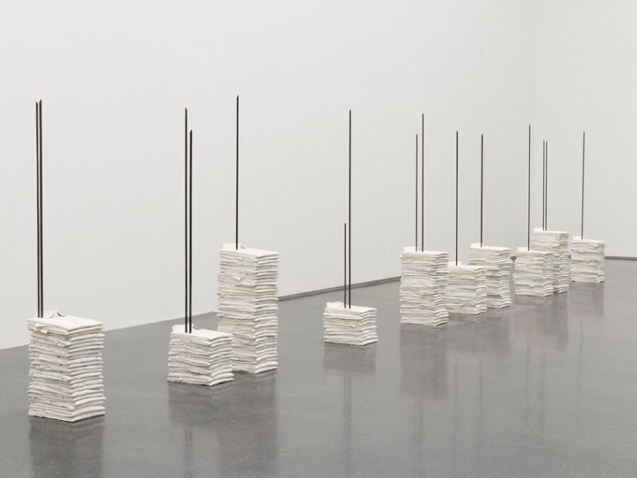 Eight piles of white shirts of various heights with rebar pierced through them in a row in a white gallery space