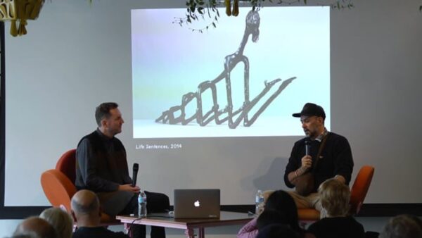 Two people sit on opposite sides of a projection screen displaying an image of a wiry sculpture