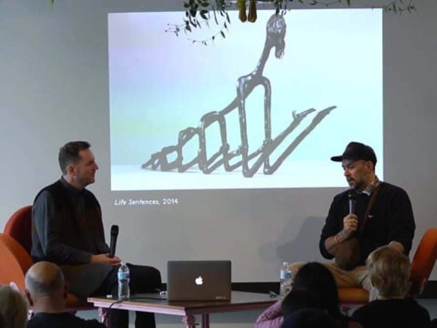 Two people sit on opposite sides of a projection screen displaying an image of a wiry sculpture