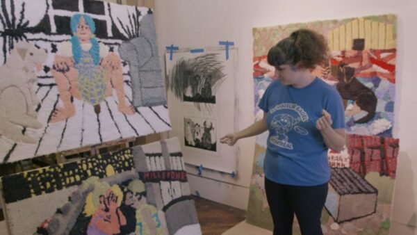 A female-presenting person in a blue shirt with curly brown hair pulled up gestures to an artwork on an easel in a studio space with other artworks hanging on the wall and leaning against things on the floor