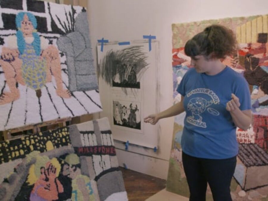 A female-presenting person in a blue shirt with curly brown hair pulled up gestures to an artwork on an easel in a studio space with other artworks hanging on the wall and leaning against things on the floor