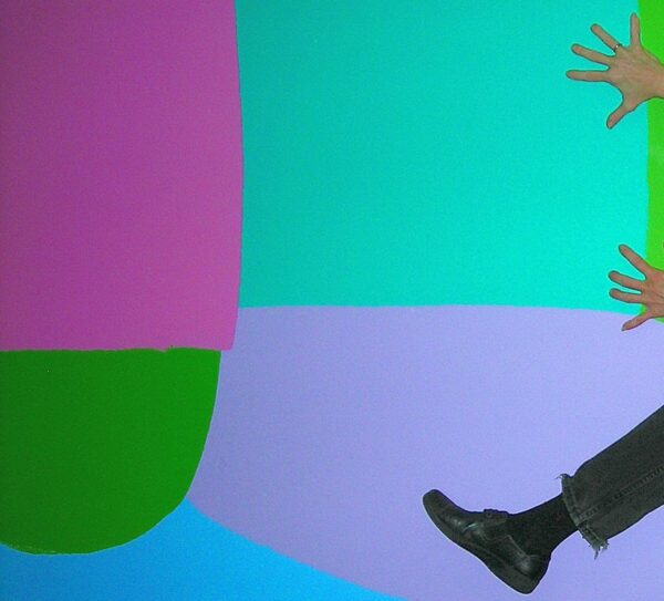 A pair of hands and the lower half of a leg just barely appear on the right-hand side of a colorful wall painted in large blocks of hot pink, mint green, lavender, turquoise, and green