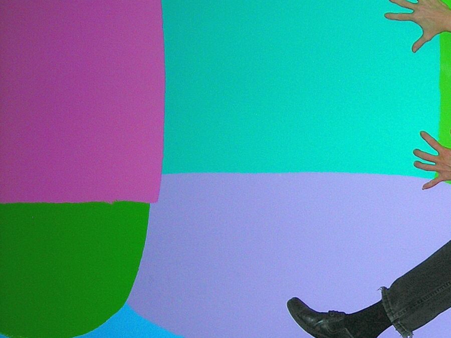 A pair of hands and the lower half of a leg just barely appear on the right-hand side of a colorful wall painted in large blocks of hot pink, mint green, lavender, turquoise, and green