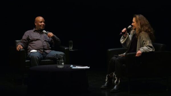 Two people holding microphones sit in black leather seats on a black stage