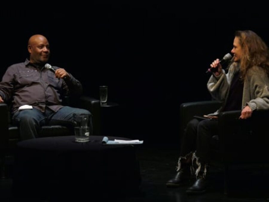 Two people holding microphones sit in black leather seats on a black stage