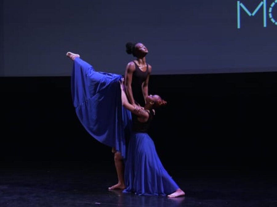 Two dancers on stage in flowy blue dresses, one lifts the other in the air