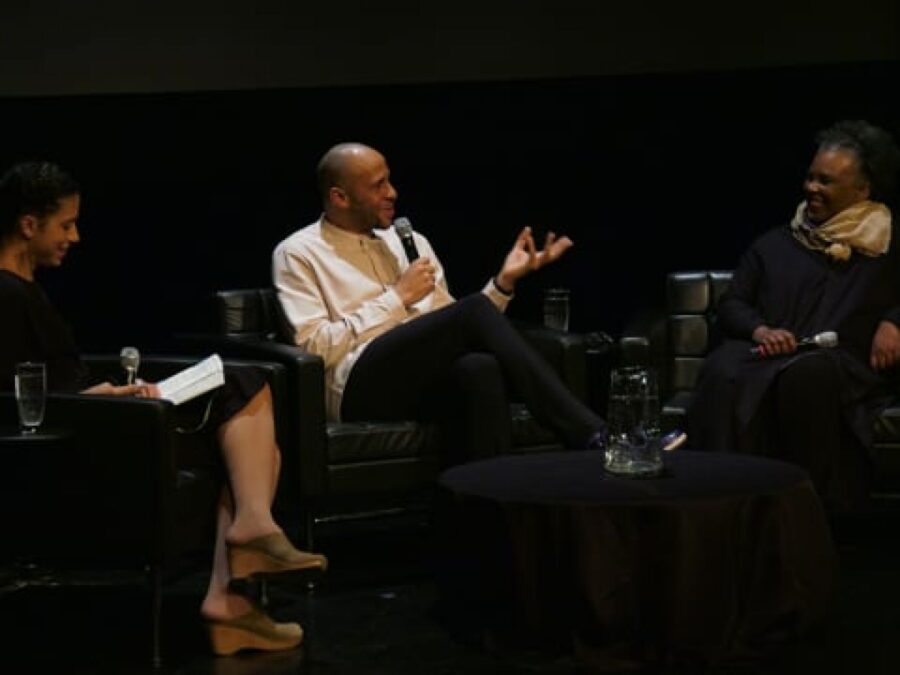 Three people sit on a dark stage looking at the person in the middle who holds a microphone up to their mouth