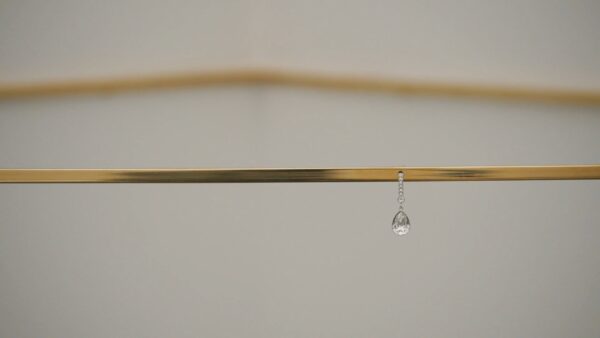 A close up image of a bronze-colored strip of metal cutting horizontally across the frame, with a single diamond earring dangling from its center. Another bent piece of similar metal is shown out of focus above and behind it.
