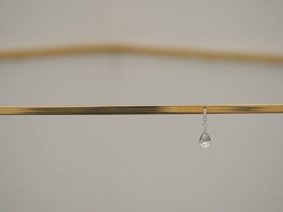 A close up image of a bronze-colored strip of metal cutting horizontally across the frame, with a single diamond earring dangling from its center. Another bent piece of similar metal is shown out of focus above and behind it.