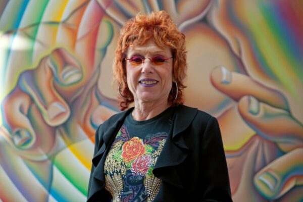 Judy Chicago, a woman with pink hair and pink glasses, poses in front of a colorful mural