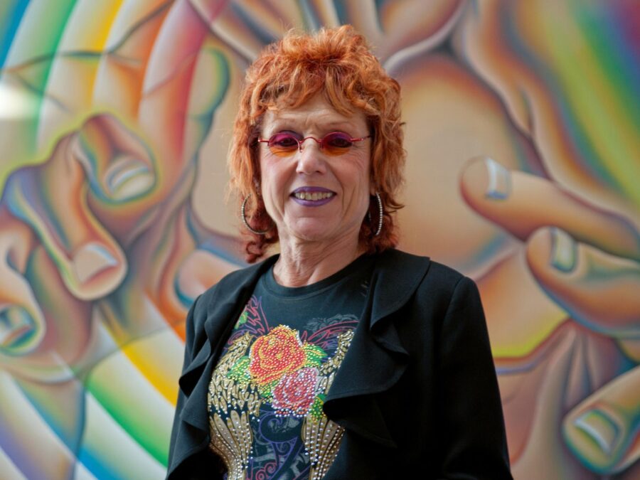 Judy Chicago, a woman with pink hair and pink glasses, poses in front of a colorful mural