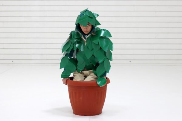 A person covered in plastic green leaves crouches in a plastic planter pot in a room with a white brick wall