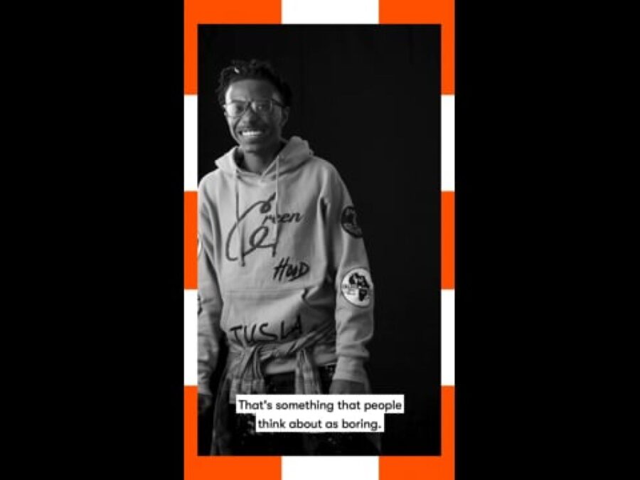 A vertical black and white video still of a male-presenting person standing with a caption at the bottom reading That's something that people think about as boring framed in orange and white dashes