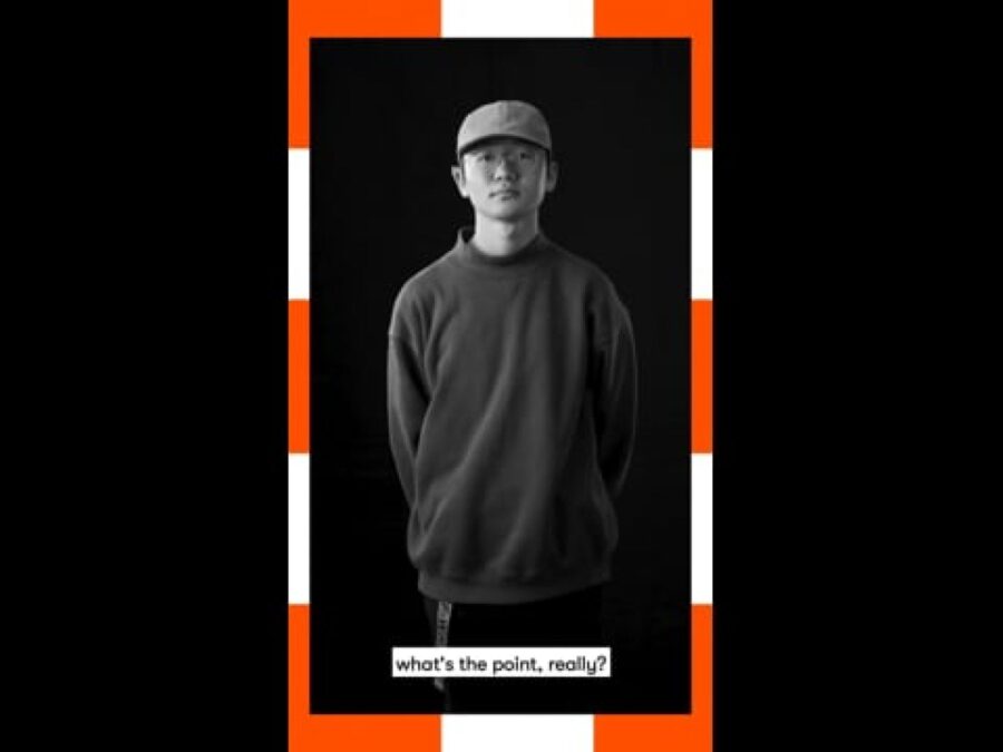 A vertical black and white video still of a male-presenting person standing with a caption at the bottom reading What's the point really, framed in orange and white dashes