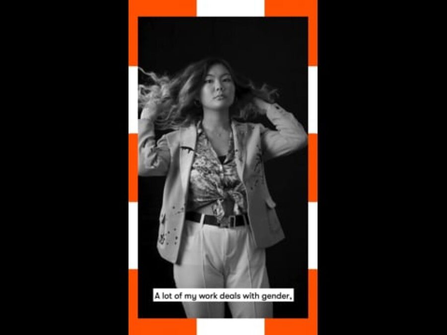 A vertical black and white video still of a female-presenting person standing flicking their hair back with a caption at the bottom reading A lot of my work deals with gender, framed in orange and white dashes