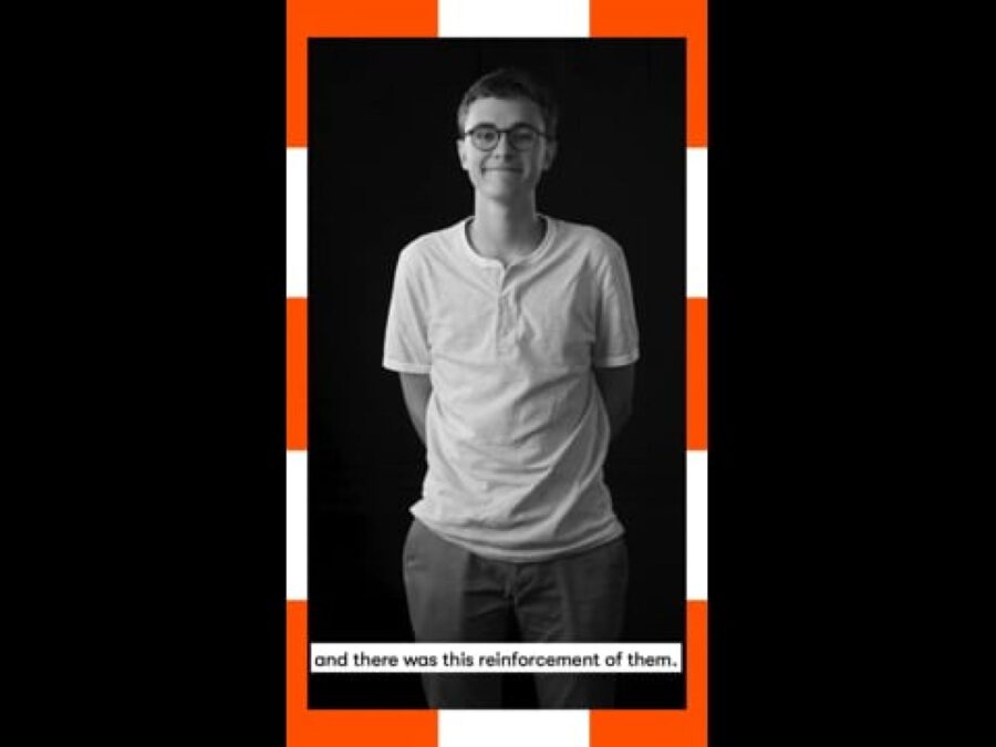 A vertical black and white video still of a male-presenting person standing with a caption at the bottom reading And there was this reinforcement of them, framed in orange and white dashes