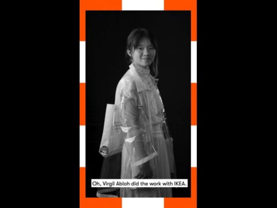 A vertical black and white video still of a female-presenting person standing with a caption at the bottom reading Oh, Virgil Abloh did the work with IKEA, framed in orange and white dashes