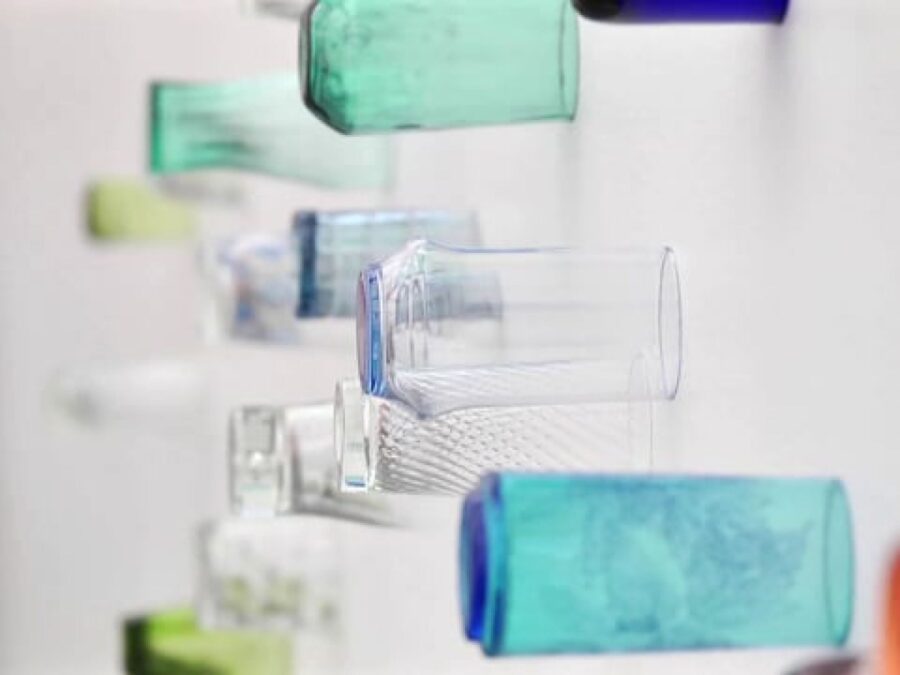 Glasses of all shapes and sizes in greens, blues, clear, and oranges attached to a white wall