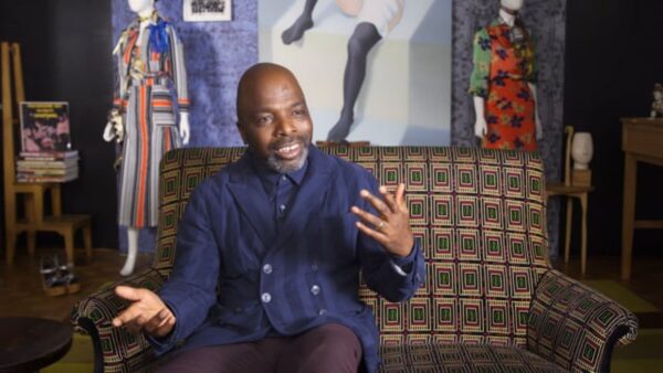 Duro Olowu gestures with his hands on a patterned couch amid artwork