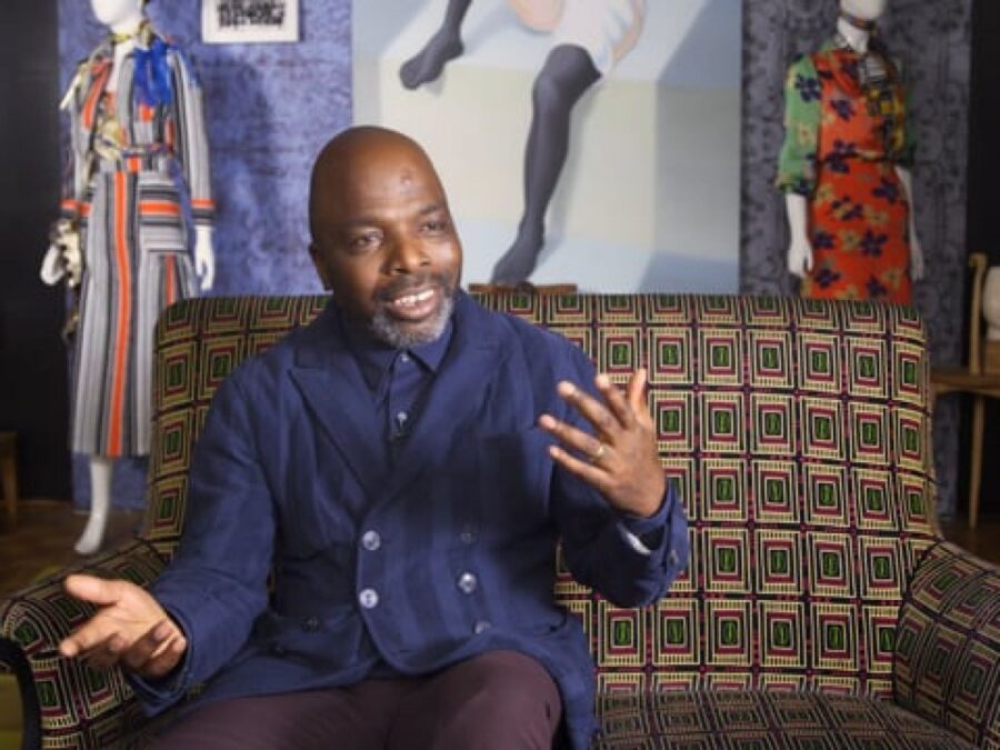 Duro Olowu gestures with his hands on a patterned couch amid artwork