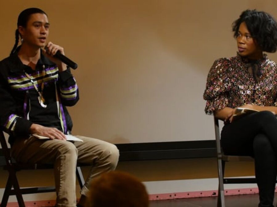 Two people sit on a stage in folding chairs as one holds a microphone up to their mouth