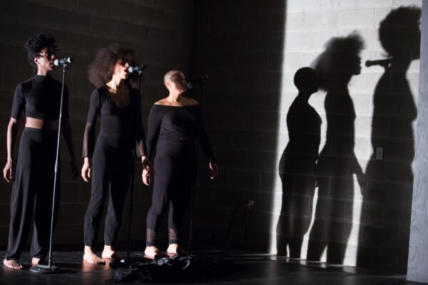 Three people in all black stand behind three microphones, their shadows cast on the wall to their lefts by a bright white light, lending a very stark contrast to the image