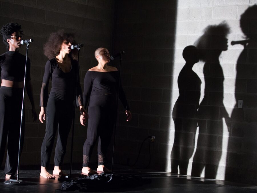 Three people in all black stand behind three microphones, their shadows cast on the wall to their lefts by a bright white light, lending a very stark contrast to the image