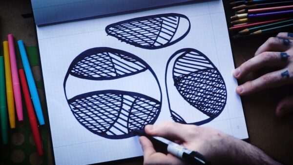 A white piece of paper with three black circular shapes on it held in place by somebody's right hand as the left hand holds a Sharpie marker to the page to finish one of the circular outlines. Bright markers and colored pencils appear on either side of the paper.