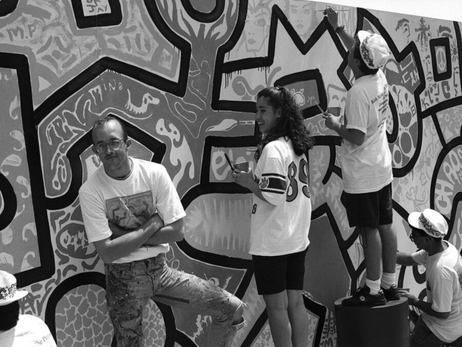 Teenagers in shorts paint a mural of graphic designs as a young man in glasses stands with arms folded looking at the camera.