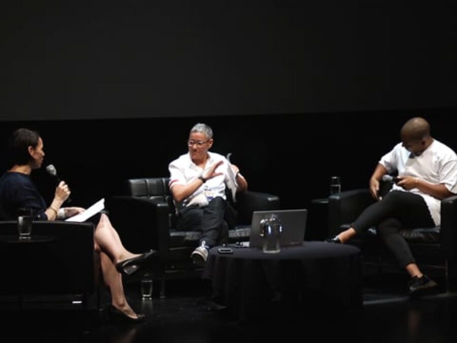 Three people sit in chairs situated around three sides of a coffee table on a black stage