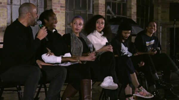 Six people sit in a row in an old brick building. Most look towards the closest person who holds a microphone while they laugh