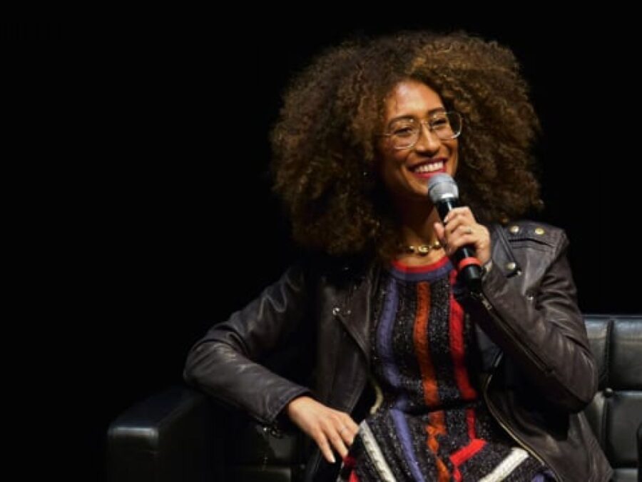 A person with full brown hair and milk chocolate skin sits on a black stage while they hold a microphone to their mouth with their left hand
