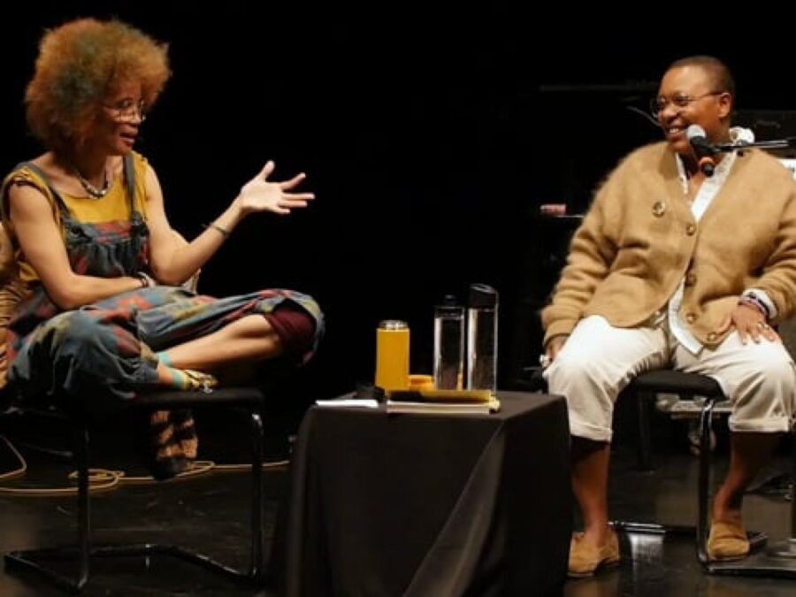 Two people sitting on a black stage with a table between them