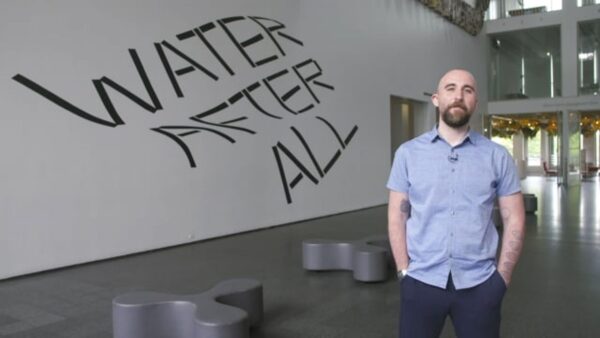 A person with a brown beard, light skin and short hair stands with their hands in their pockets in front of wavy text reading Water After All