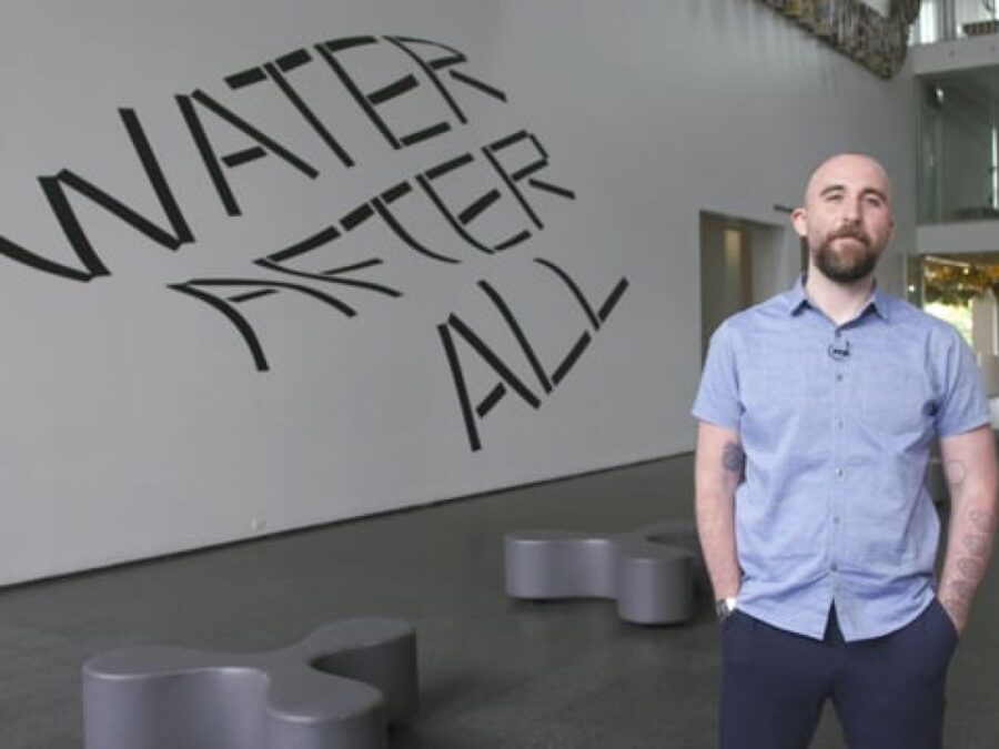 A person with a brown beard, light skin and short hair stands with their hands in their pockets in front of wavy text reading Water After All