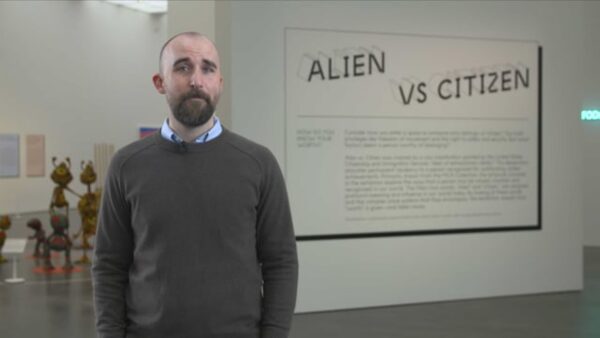 A person with a beard and shortly trimmed brown hair stands near large text reading Alien vs Citizen with artwork in the background