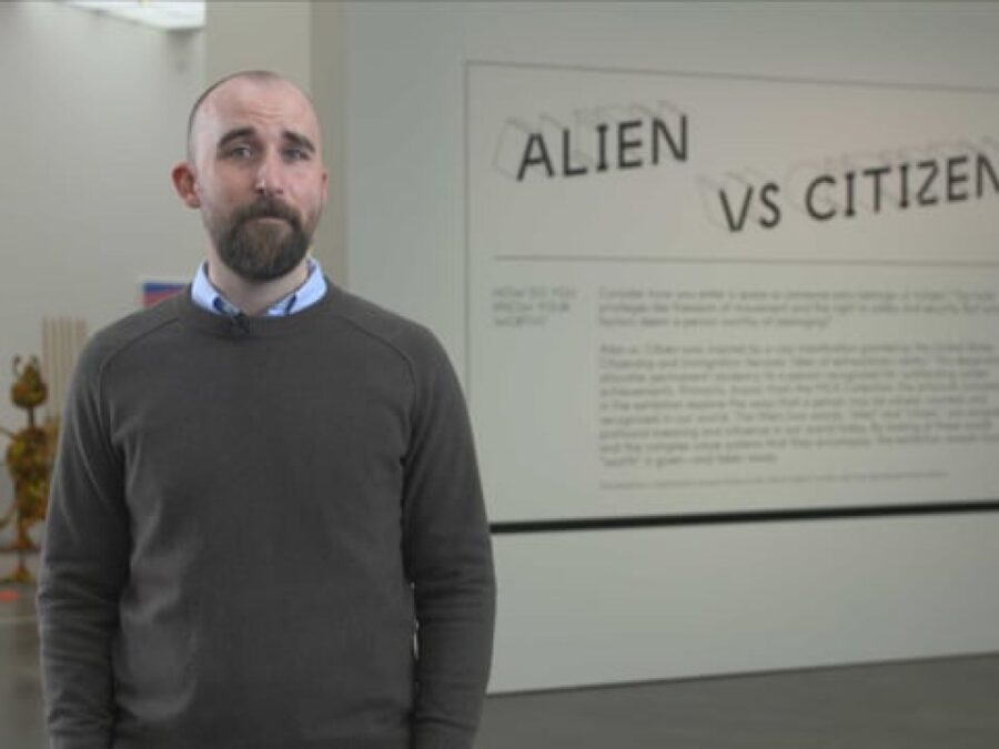 A person with a beard and shortly trimmed brown hair stands near large text reading Alien vs Citizen with artwork in the background