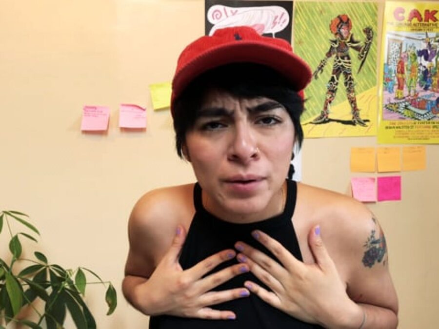 A person with both hands on their chest looks directly at the camera with a confused look on their face. Behind them are post-it notes, posters and a plant