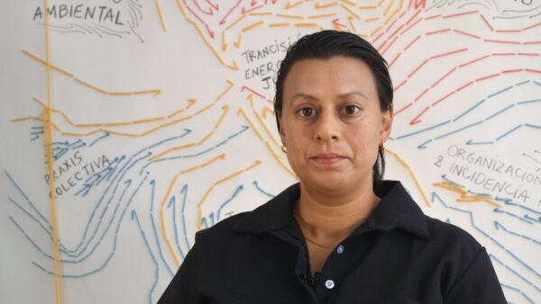 A person with tightly pulled back hair poses in front of a wall filled with writing and blue and orange arrows